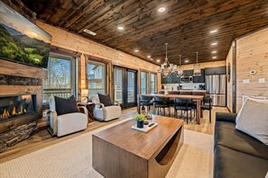 A modern log cabin interior with a fireplace, dining area, kitchen, and seating. - Cabin w/ Rooftop Bar · Indoor Pool · Mtn View (Pigeon Forge)