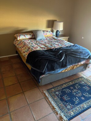 Un dormitorio con cama, mesita de noche, lámpara, alfombra azul y colcha estampada.