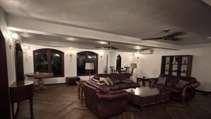 Ein geräumiges Wohnzimmer mit Fischgrätparkett, einer braunen Ledersitzgruppe, einem Couchtisch, einem Esstisch mit Stühlen und einem Bücherregal.