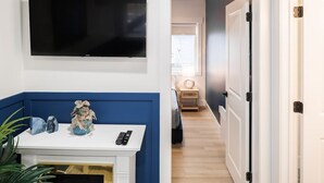Un couloir avec un téléviseur fixé au mur, une console blanche avec une télécommande, et une porte donnant sur une autre pièce.