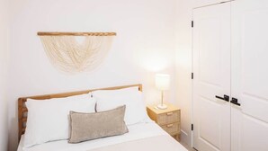 Een bed met witte beddengoed, een houten hoofdbord, een beige kussen, een bedlampje, een geweven wanddecoratie en een witte kledingkast.