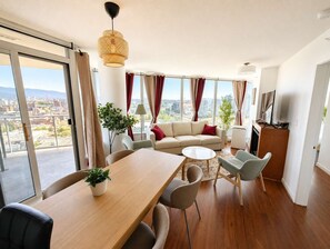A modern living room with a sofa, coffee table, and dining area, large windows with curtains, and a city view. - Luxe Retreat | BCPlace/Skytrain/View/Pool/Jacuzzi (Vancouver)