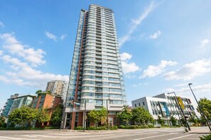A modern high-rise building with a curved facade, surrounded by other contemporary buildings and greenery. - Luxe Retreat | BCPlace/Skytrain/View/Pool/Jacuzzi (Vancouver)