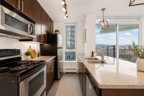 A modern kitchen with a stove, microwave, refrigerator, and a large window offering a city view. - Luxe Retreat | BCPlace/Skytrain/View/Pool/Jacuzzi (Vancouver)