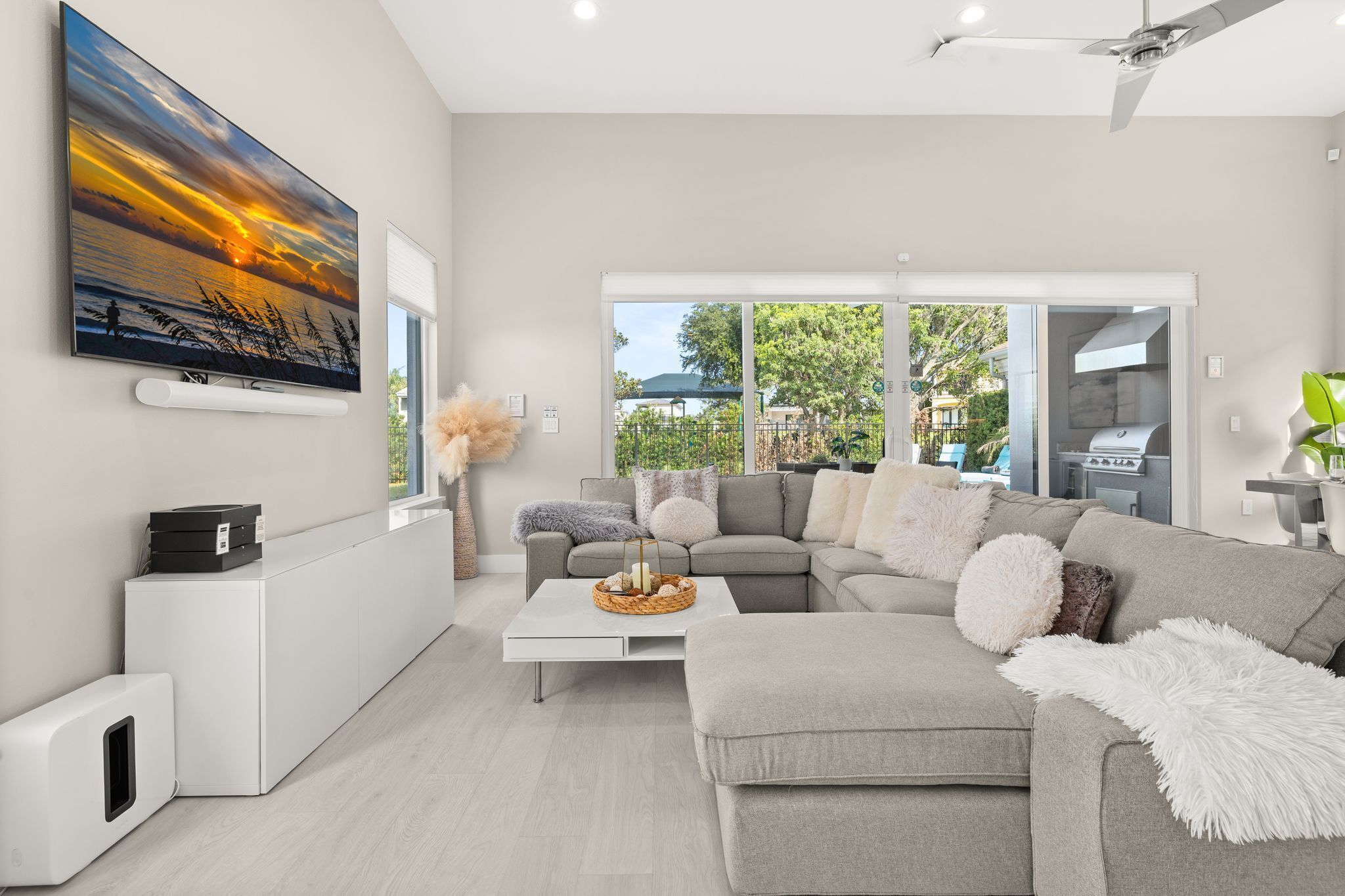 A modern living room with a large flat-screen TV, a grey sectional sofa, a white coffee table, and a view of the outdoors through large windows.