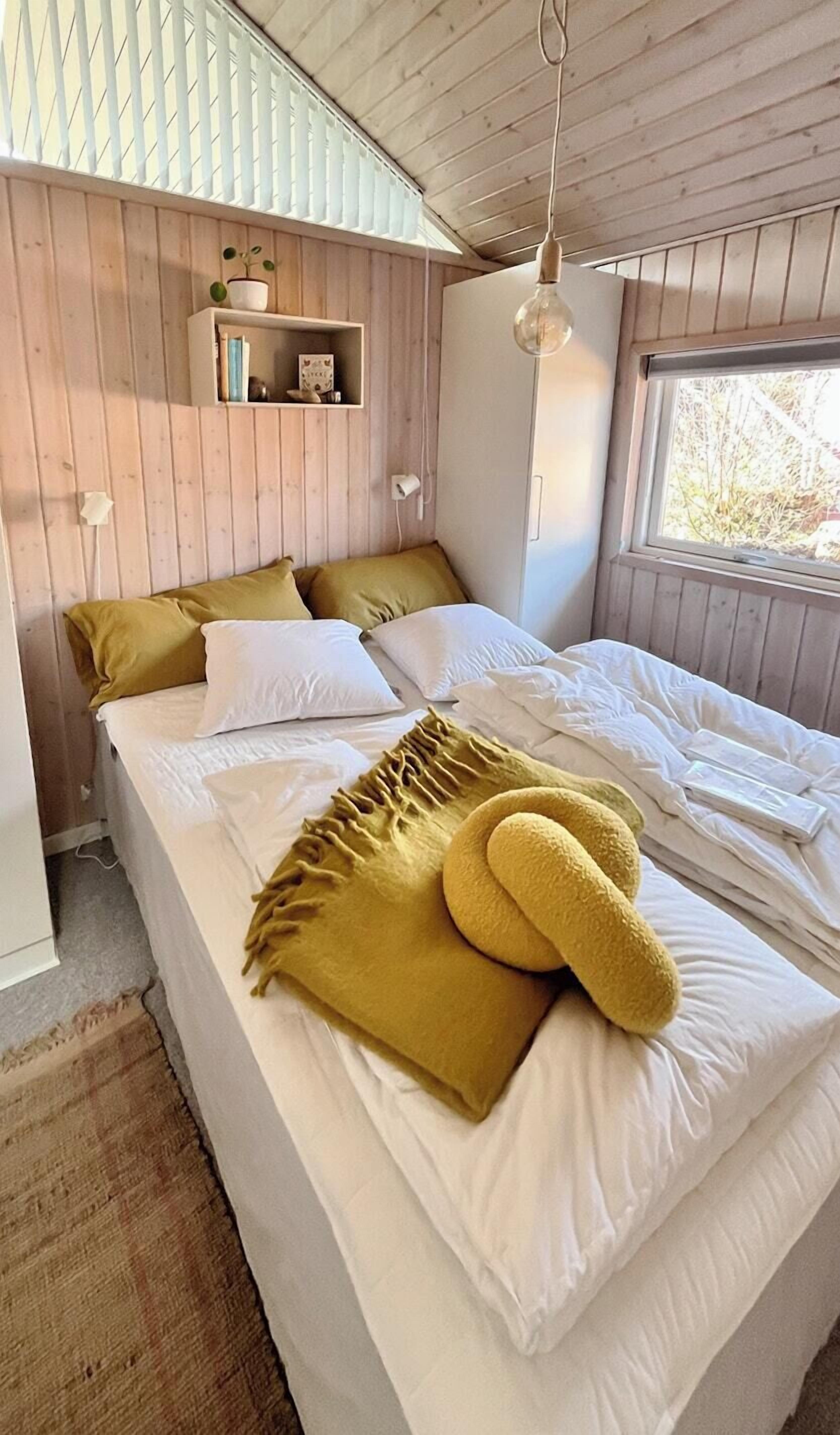 Un dormitorio con cama, una estantería con libros y una planta, además de un ventanal.