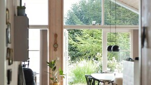 Een smalle gang met een hoge plafond, houten balken en grote ramen. Er is een eethoek met een tafel en stoelen, en een potplant.