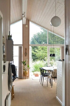 A narrow hallway with a high ceiling, wooden beams, and large windows. There is a dining area with a table and chairs, and a potted plant. - Summerhouse by the Little Belt with sea view (Haderslev)