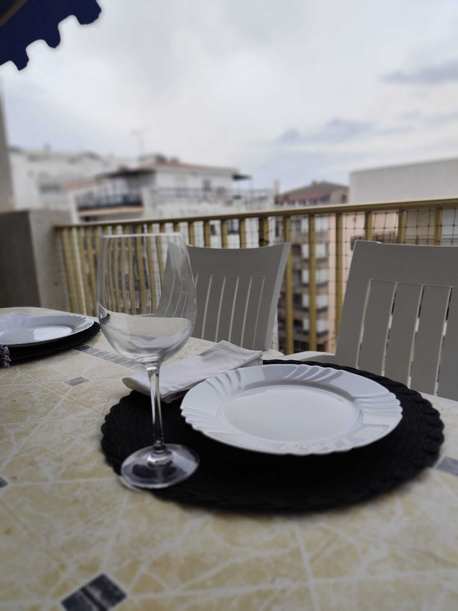 Una mesa dispuesta con platos, una copa de vino, sillas y una vista de edificios al fondo.