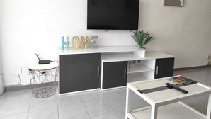 Een moderne woonkamer met een flatscreen televisie aan de muur, een witte salontafel, een klein rond tafeltje met een stoel en een plant in pot.