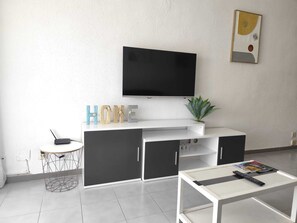 A modern living room with a flat-screen TV mounted on the wall, a white coffee table, a small round table with a chair, and a potted plant. - Pigem Apartment (Salou)