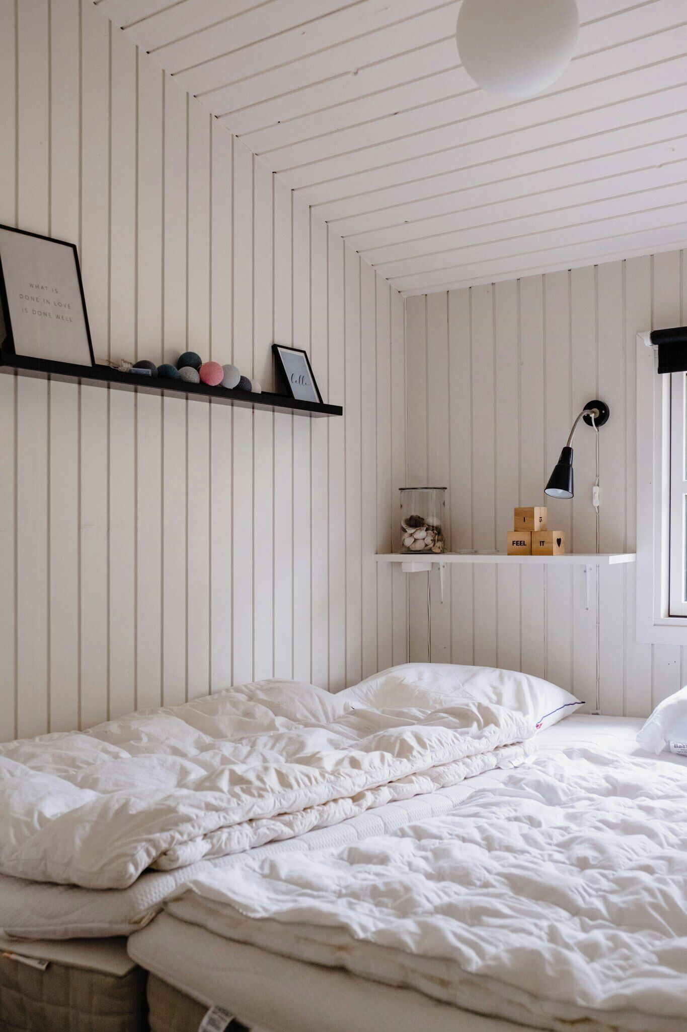 A bedroom with a bed, white walls, a shelf with decorative items, and a bedside lamp.