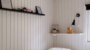 A bedroom with a bed, white walls, a shelf with decorative items, and a bedside lamp.