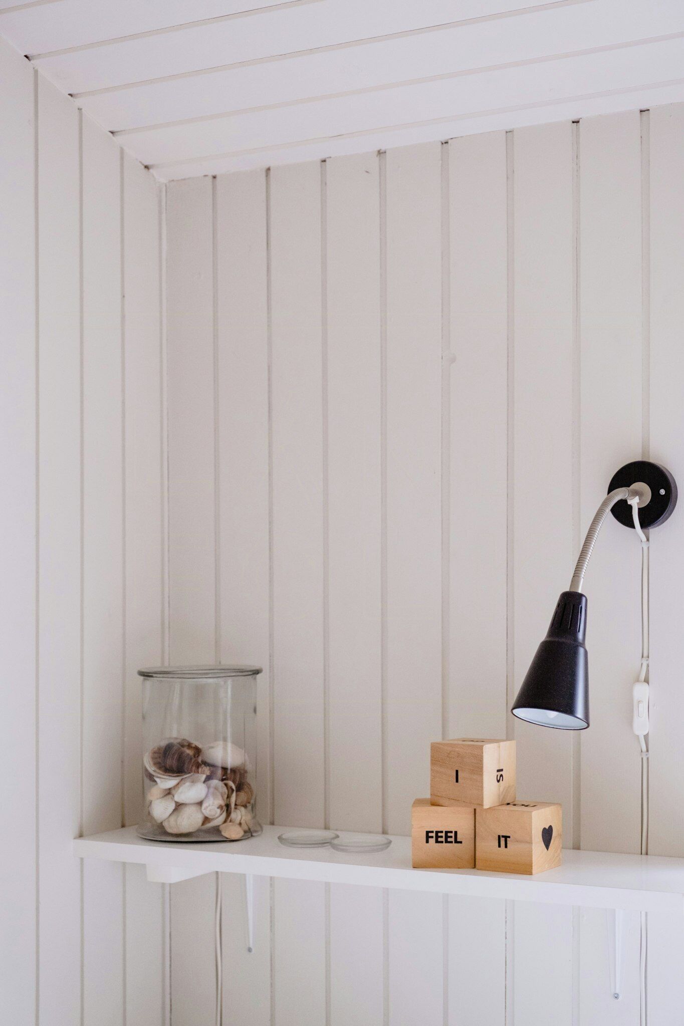 A white shelf with a glass jar of seashells, wooden blocks spelling 'I FEEL IT,' and a black desk lamp.