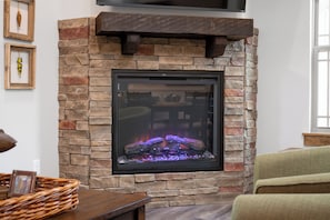 Una chimenea de piedra con un televisor montado encima, una cesta de mimbre sobre una mesa, y un sillón verde en primer plano.
