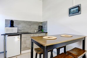 A compact kitchen with a microwave, coffee maker, and a small refrigerator. There is a wooden table with two stools and a wall-mounted power outlet. - Liyann - Proche plage - Terre Sainte (Saint-Pierre)