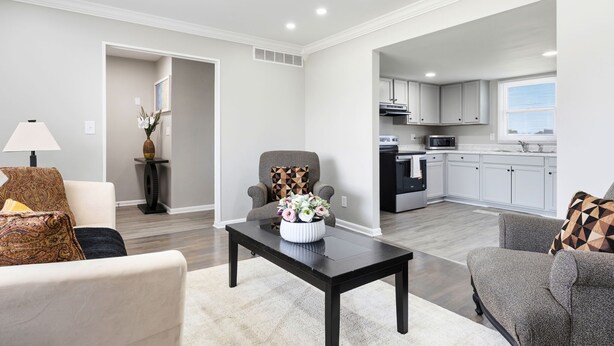 Un moderno salón con sofá, sillón y mesa de centro, comunicado a una cocina con armarios blancos y una ventana.