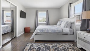 Un dormitorio con cama, televisor, mesita de noche, alfombra y ventana con cortinas.