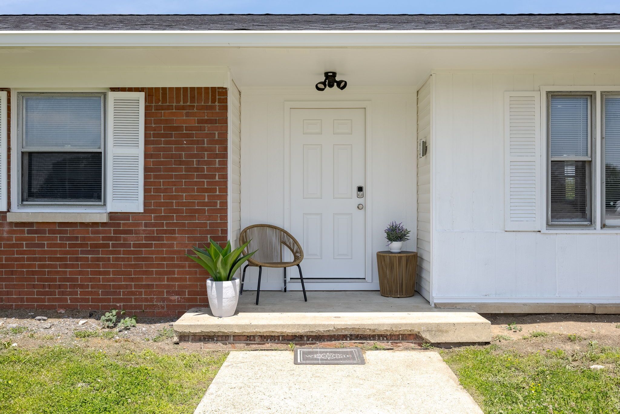 A house with a white front door, a brick wall, a potted plant, a wicker chair, and a small round table.