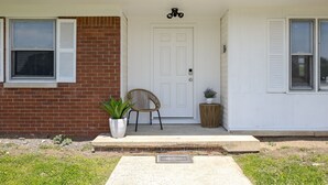 Una casa con puerta principal blanca, pared de ladrillo, planta en maceta, silla de mimbre y una pequeña mesa redonda.