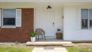 A house with a white front door, a brick wall, a potted plant, a wicker chair, and a small round table.