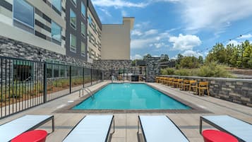 A rectangular outdoor swimming pool with a tiled deck, surrounded by a stone wall and a metal fence. There are lounge chairs and a bar area with yellow chairs.