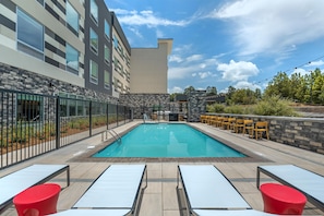 A rectangular outdoor swimming pool with a tiled deck, surrounded by a stone wall and a metal fence. There are lounge chairs and a bar area with yellow chairs.