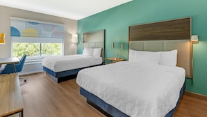 A hotel room with two beds, a desk, and a window with a view of trees.