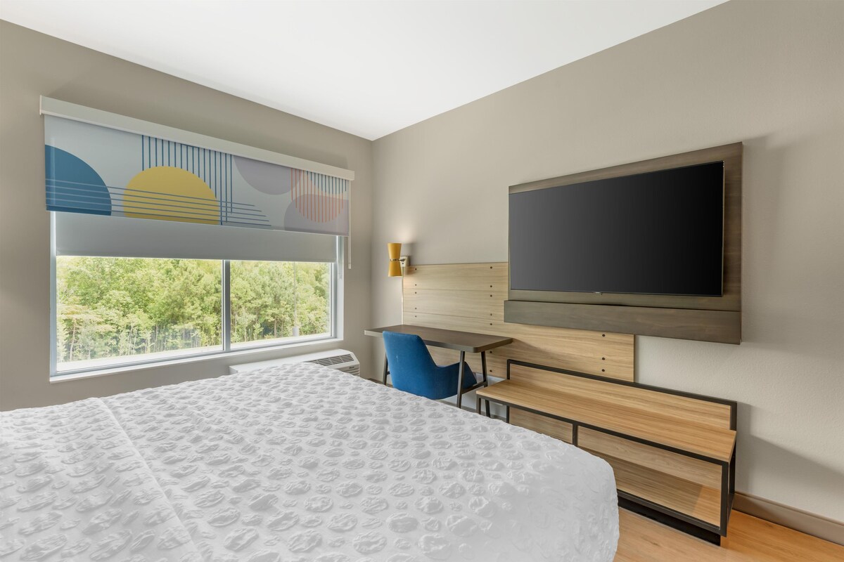 A hotel room with a bed, a desk, a chair, a TV, and a window with a view of trees.