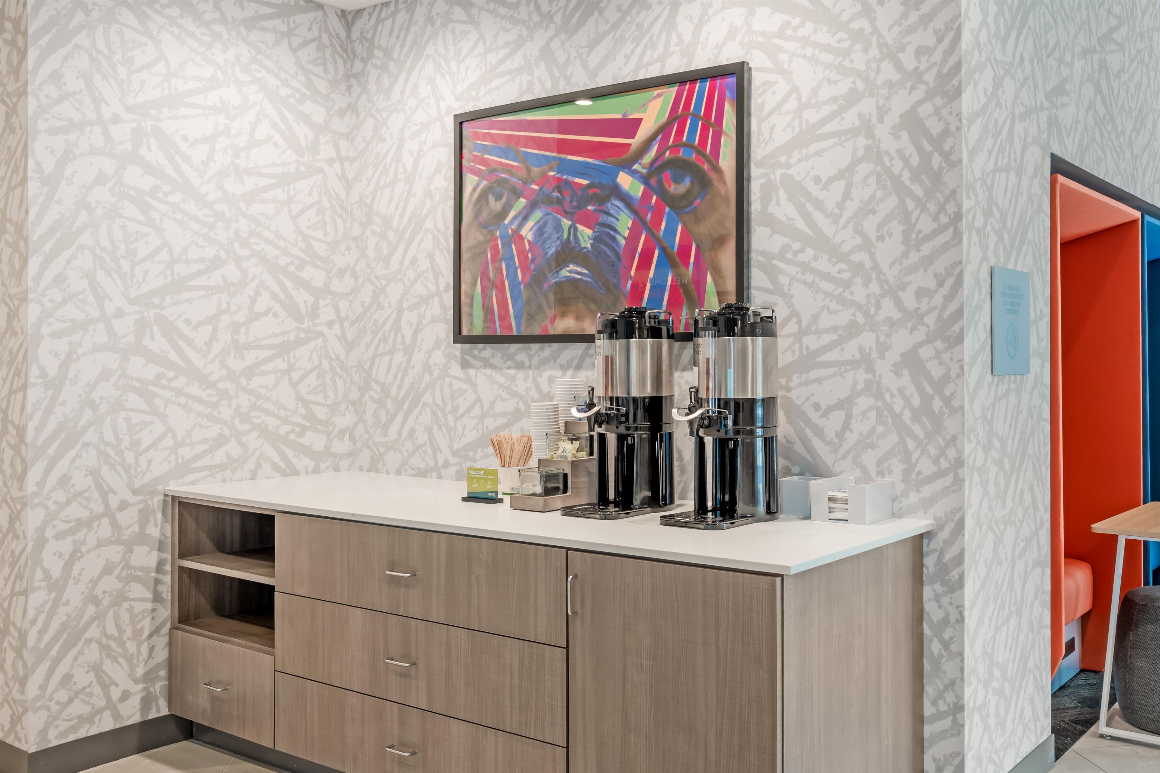A modern hotel room with a white counter, three coffee machines, a welcome sign, and a colorful abstract painting on the wall.
