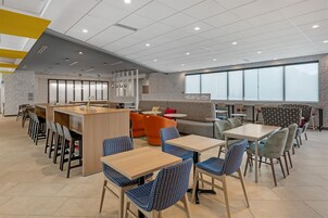 Un café moderno con zona de bar, diferentes disposiciones de asientos y amplios ventanales.