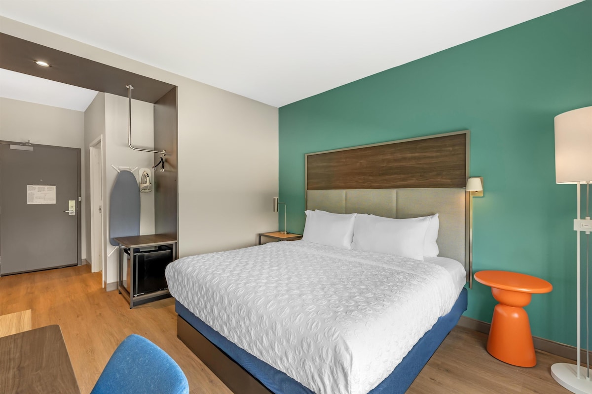 Una habitación de hotel con cama, escritorio, silla, mesita, lámpara y baño.