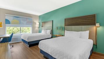 A hotel room with two beds, a desk, and a window with a view of trees.