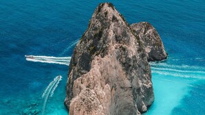 A rocky island surrounded by clear turquoise waters, a boat creating a wake, and a sandy beach. - Villy Suites II - Zante Lux Suite Near the Beach (Zakynthos)
