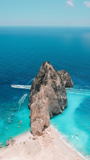 Eine felsige Insel, umgeben von klarem türkisblauem Wasser, ein Boot zieht eine Schneise und ein Sandstrand.