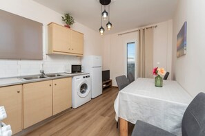 Eine kompakte Küche mit Waschmaschine, Mikrowelle und Kühlschrank. Ein Essbereich mit Tisch und Stühlen lädt zum Verweilen ein, und eine kleine Pflanze ziert die Wand.