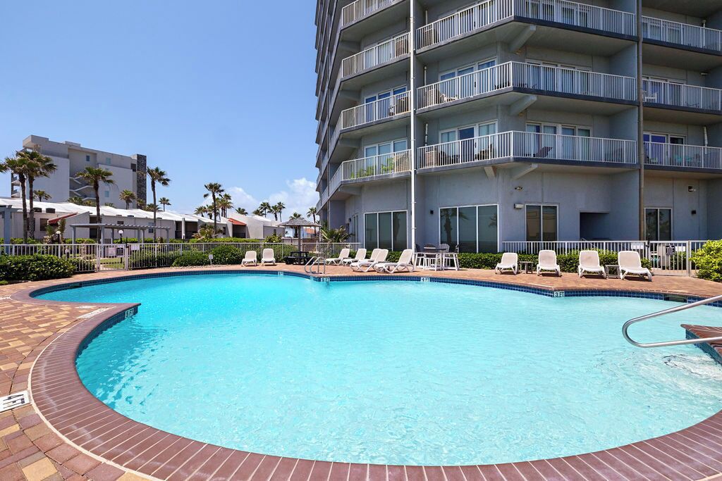 A swimming pool with lounge chairs, a multi-story building with balconies, and palm trees in the background.