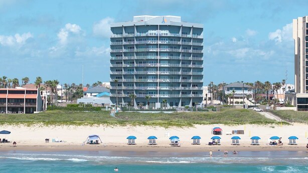 Een hotel met meerdere verdiepingen en balkons, nabij een strand met parasols en een zandstrand.