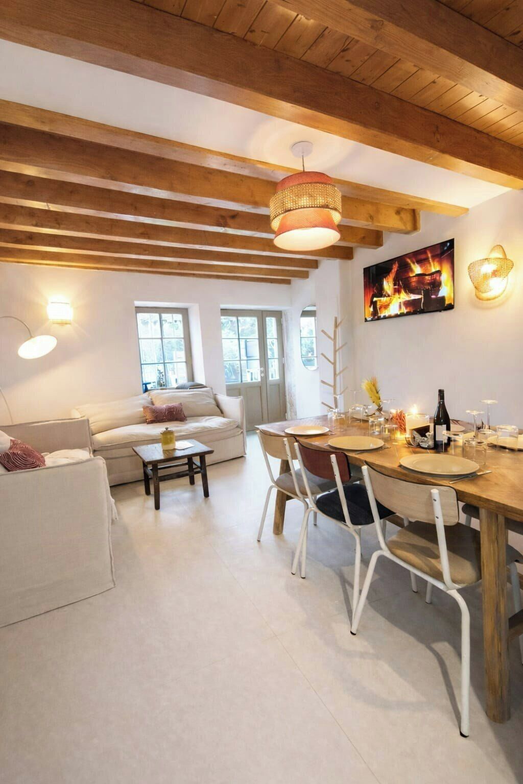 A cozy living room with a wooden ceiling, a sofa, a dining table set for four, and a fireplace with a painting above it.