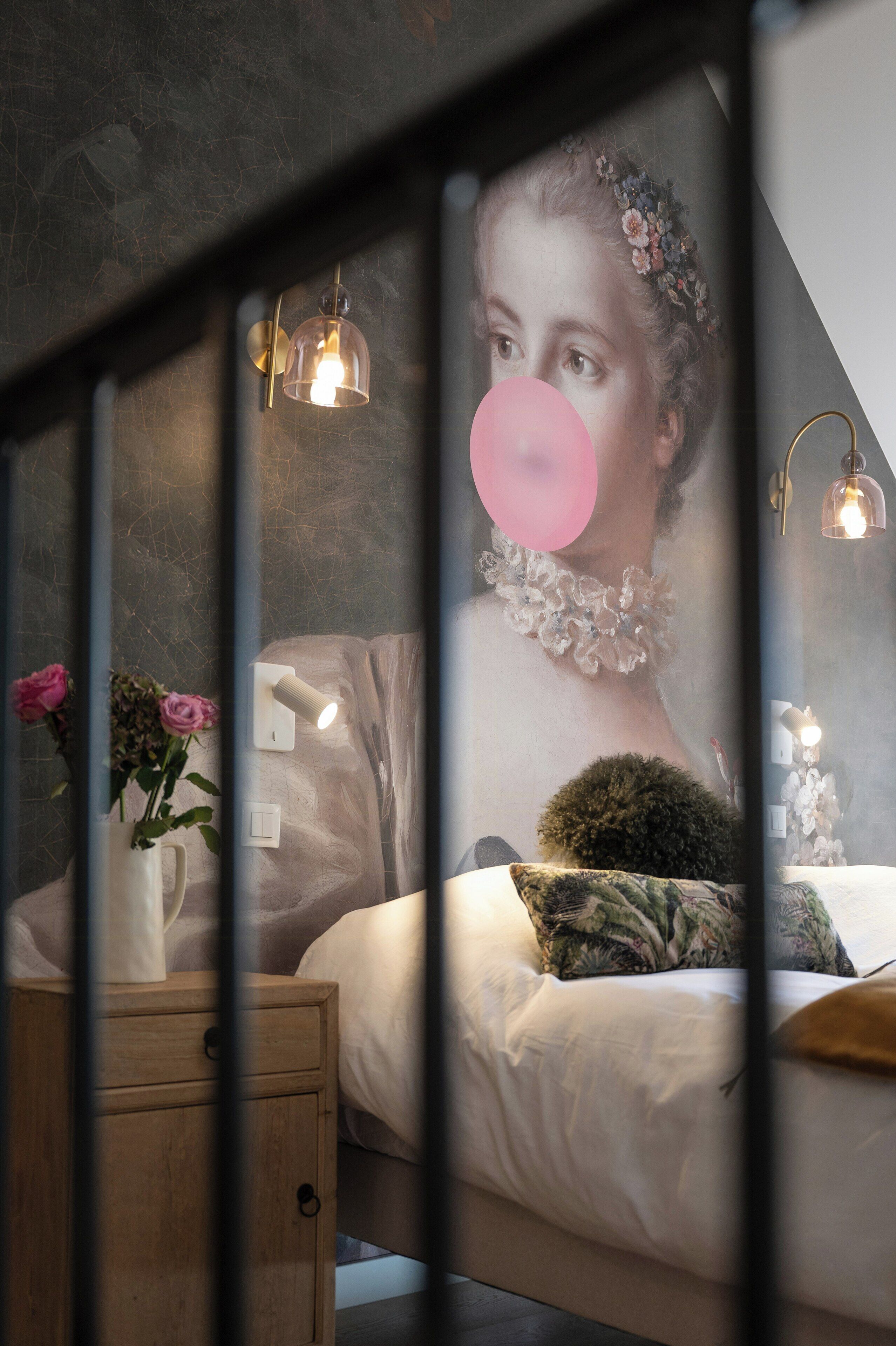 Une chambre à coucher avec un lit, une table de chevet, un vase de fleurs et un mur orné d’un motif floral.