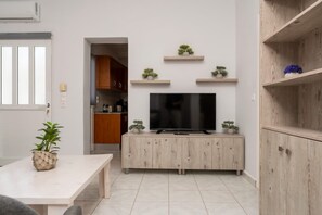 Ein modernes Wohnzimmer mit Fernseher auf einer Holzkommode, einem Esstisch mit Pflanze und einer Küche im Hintergrund.