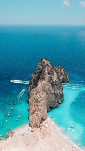 A rocky island surrounded by clear turquoise waters, a boat creating a wake, and a sandy beach. - Villy Suites I - Zante Luxury Suite With Hot Tub (Zakynthos)