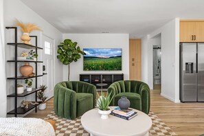 Ein modernes Wohnzimmer mit einem großen Flachbildfernseher, zwei grünen Sesseln, einem runden Couchtisch und einem Bücherregal mit Dekoration.