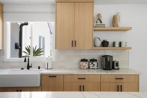 Eine moderne Küche mit Holzschränken, einer weißen Arbeitsplatte, einer Spüle, einer Kaffeemaschine und einem Fenster mit Blick auf Gebäude.
