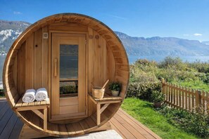 Eine Holzsauna mit Tür, zwei Handtüchern, einem Eimer und einer Pflanze auf einer Terrasse mit Blick auf Berge und See.