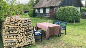 Un área de estar al aire libre rústica, con una mesa, sillas, una pila de leña y una casa con techo de paja al fondo.