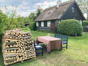 Ein rustikaler Außenbereich mit Tisch und Stühlen, einem Holzstapel und einem Haus mit Strohdach im Hintergrund.