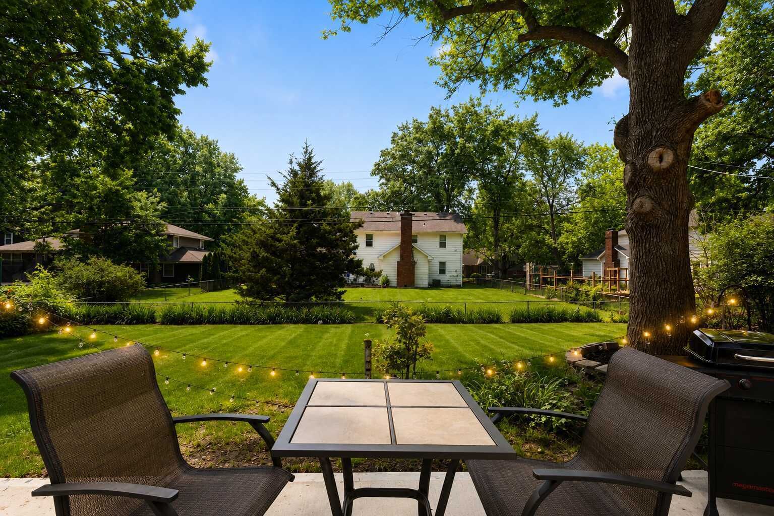 An outdoor patio with a table and chairs, surrounded by trees and a well-maintained lawn.