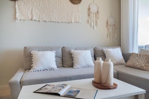 Ein Wohnzimmer mit grauem Sofa, weißen Kissen, einem weißen Couchtisch und Dekorationselementen wie Wandbehängen aus Makramee und Traumfängern.