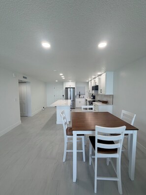 A modern kitchen with white cabinets, a stainless steel refrigerator, and a dining area with a wooden table and white chairs. - Shores Club 1004 (Daytona Beach Shores)